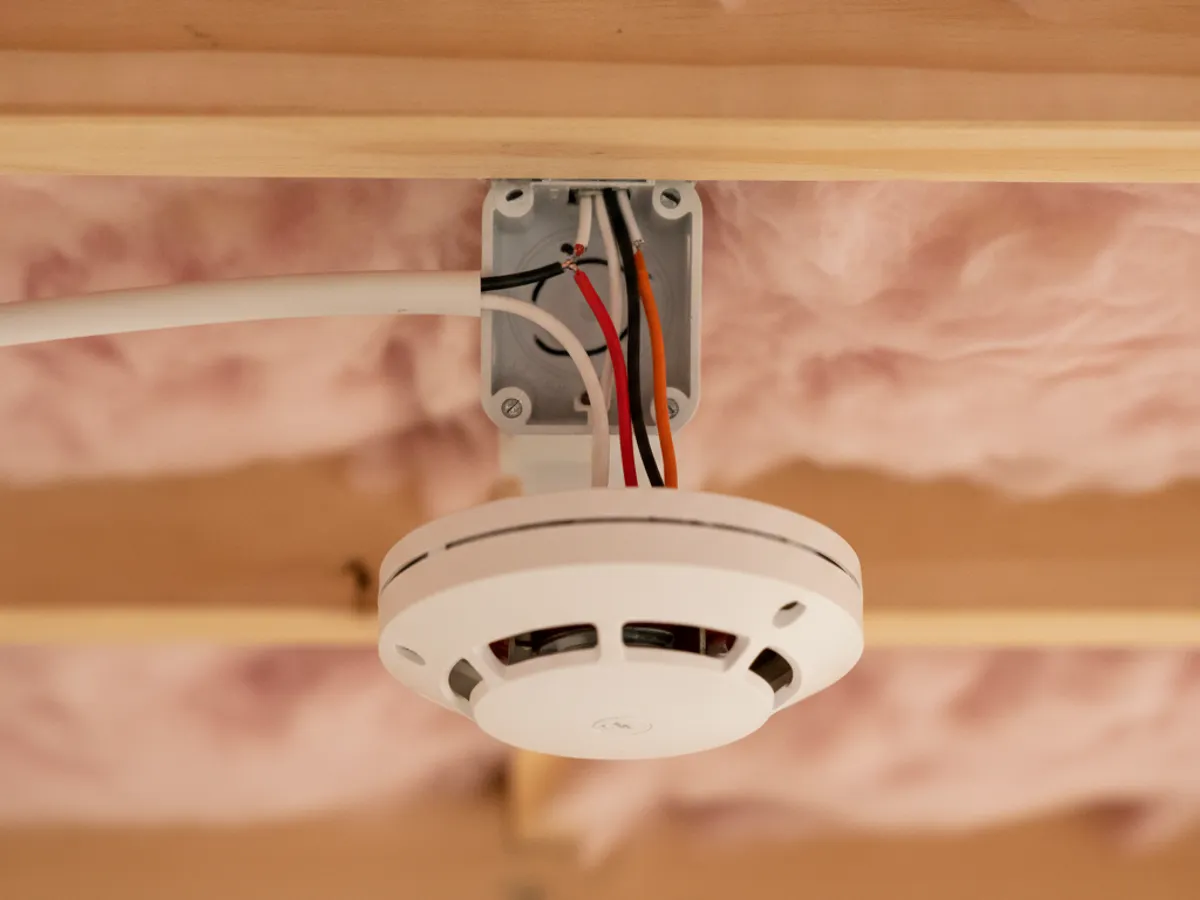 Close-up of a hardwired smoke detector base with an interconnect wire and wire nuts inside an open ceiling cavity